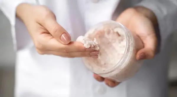 Dłonie w białym kitlu pobierają z pojemnika drobnoziarnisty peeling. To świetny sposób, by dowiedzieć się, co to mocznik i jak działa na skórę.
