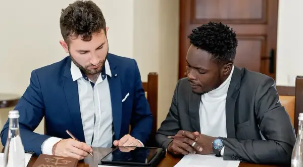 Dwóch mężczyzn w garniturach przy stole, jeden pisze, drugi patrzy. Rozmawiają o tym, jak zwracamy się do notariusza.