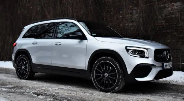 Srebrny Mercedes GLB 200 na czarnych felgach, zaparkowany na zaśnieżonej drodze przy ceglanym murze.