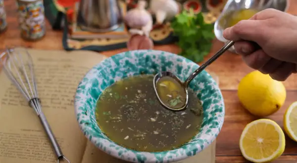 Un mestolo raccoglie il salmoriglio da una ciotola, pronto per la ricetta. Ingredienti freschi e un libro di cucina completano la scena.