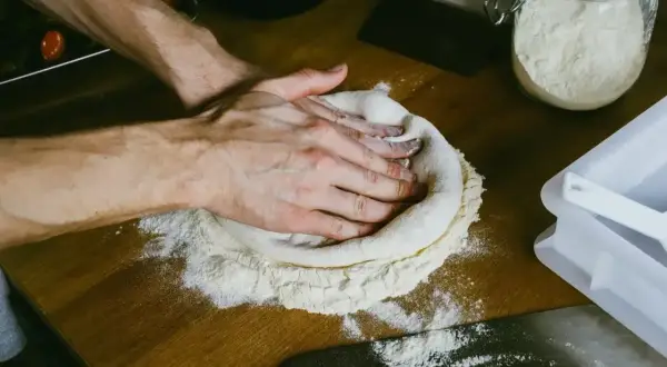 Dłonie formują ciasto na pizzę na drewnianym blacie. W tle słoik z mąką i sos pomidorowy. Zastanawiasz się, ile ma rosnąć ciasto na pizzę?