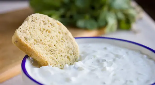 Ein Stück Brot wird in eine Schüssel mit cremigem, selbstgemachtem Zaziki getunkt. Frische Kräuter im Hintergrund.