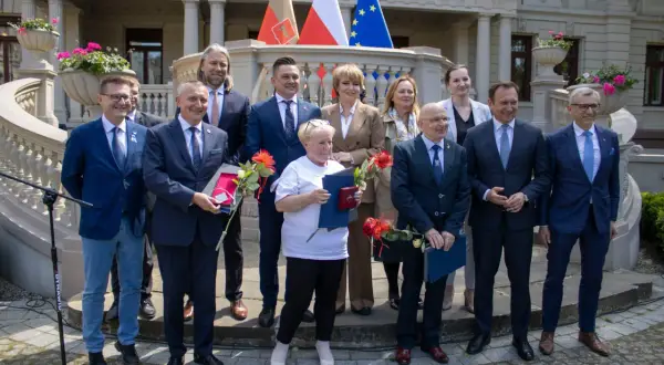Grupa osób w garniturach i strojach formalnych stoi przed budynkiem z flagami.