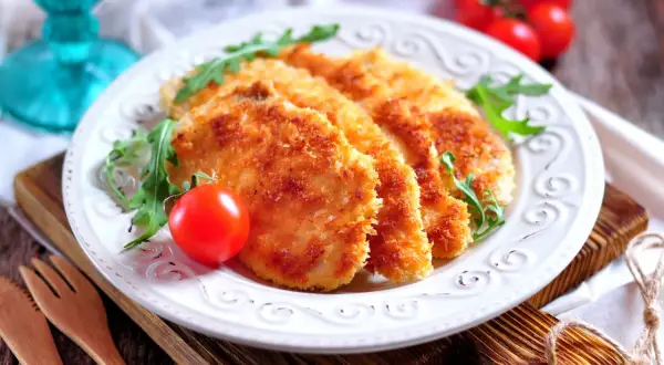 Delizioso pollo impanato al forno, croccante e dorato, servito con pomodorini e rucola su un piatto bianco.