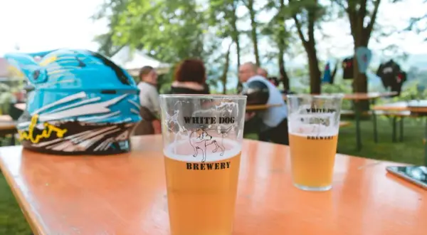 Bicchiere di birra con logo White Dog Brewery, accanto a un casco da moto blu, su un tavolo all'aperto.