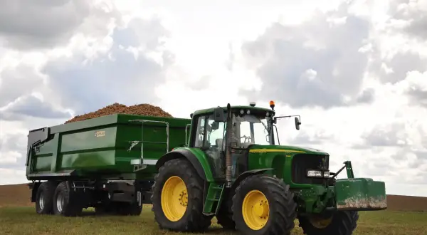 Zielony traktor John Deere z przyczepą pełną ziemniaków, wyposażoną w hamulec najazdowy, gotowy do pracy na polu.