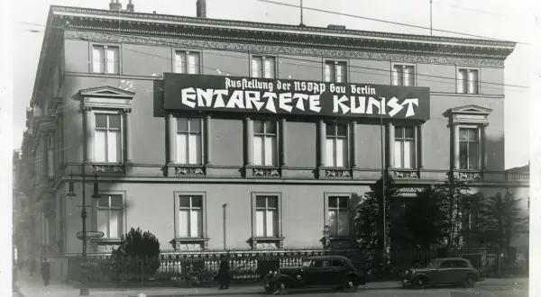 Große Banner verkünden die "Ausstellung der NSDAP Gau Berlin ENTTARTETE KUNST". Autos und Fahrräder stehen davor.