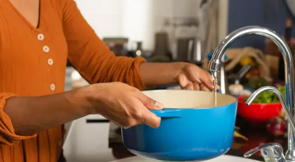 Une personne remplit une cocotte bleue sous un robinet pour faire bouillir de l'eau chaude.