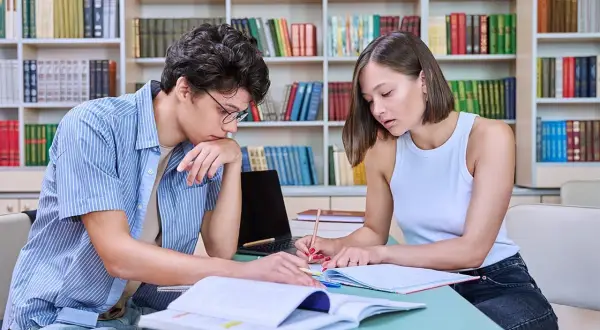 Dos personas jóvenes estudian juntas en una biblioteca, rodeadas de estanterías llenas de libros.