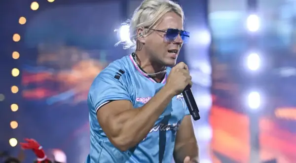 Artysta z blond włosami i niebieskimi okularami śpiewa na scenie. W tle widać światła i tłum. To koncert na **Skolim stacja**.