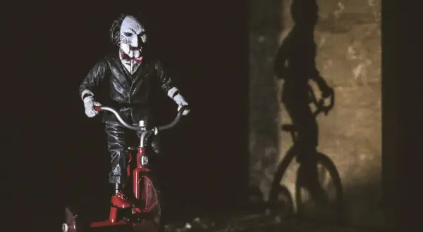 Lalka Billy z Piły na czerwonym trójkołowcu, cień na ścianie. Idealne do listy horrorów.