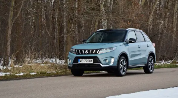 Jasnoniebieska Suzuki Vitara na drodze leśnej, z drzewami w tle i resztkami śniegu.