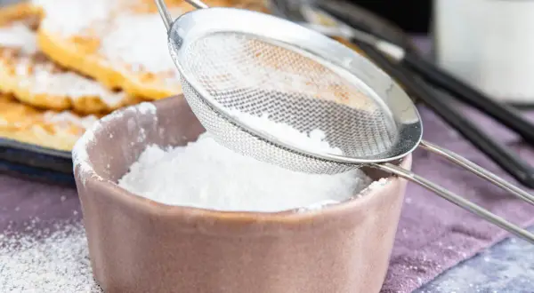 Feiner Puderzucker rieselt aus einem Sieb in eine Schale. So einfach lässt sich puderzucker selber machen für leckere Waffeln.