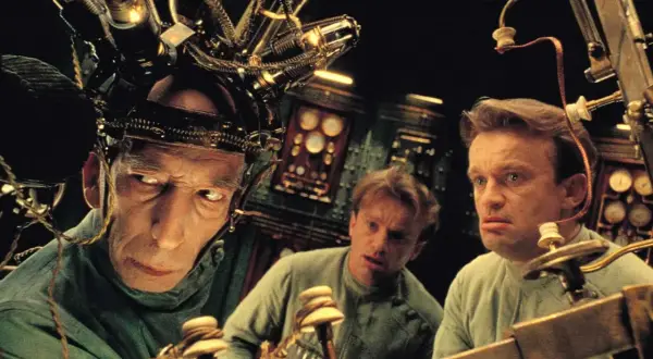 Trzej mężczyźni w dziwnych hełmach, jak z **starych filmów science fiction**, pracują przy skomplikowanej maszynie.