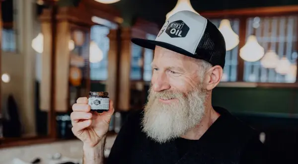 Mann mit Bart hält Tiegel "Beyer's Oil". Perfekt für Bartpflege selber machen.