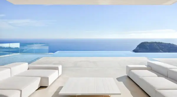 Terrasse moderne avec canapés blancs et table basse, bordée par une piscine à débordement et la mer bleue. Le **béton ciré autour de la piscine** crée une ambiance épurée et luxueuse.