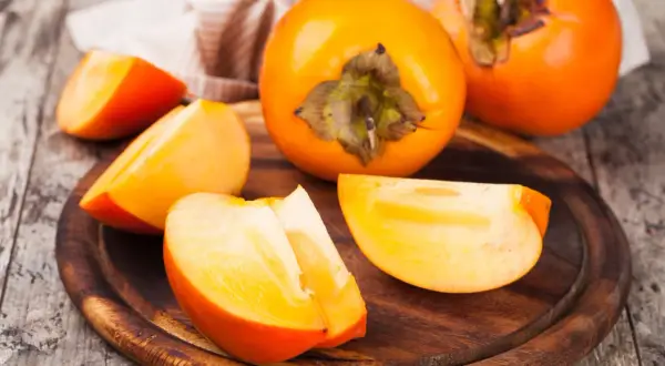 Pomarańczowe, soczyste kaki pokrojone w ćwiartki i całe owoce leżą na drewnianej desce.