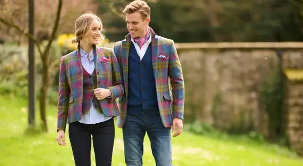 Ein Paar in farbenfrohen Tweed-Jacken und Westen spaziert durch einen sonnigen Park. Was ist Tweed? Ein robuster, traditioneller Stoff.