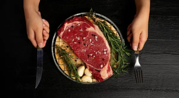 Ojo de bife crudo, sazonado con sal y pimienta, con romero y ajos, listo para cocinar. Manos sostienen cuchillo y tenedor.