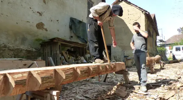 Deux hommes travaillent sur un tronc d'arbre, pratiquant l'équarrissage bois avec une hache. Le bois est façonné avec soin.