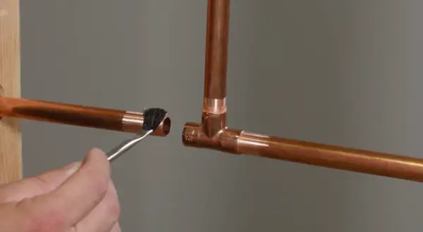 A hand applies flux with a brush to a copper pipe end, preparing it for soldering to a copper tee fitting.