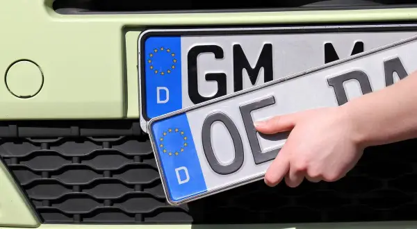 Dłoń trzyma tablice rejestracyjne państw. Jedna z nich ma oznaczenie "GM", druga "OE".