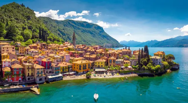 Village coloré au bord des lacs italiens, avec des montagnes verdoyantes en arrière-plan et un voilier sur l'eau turquoise.