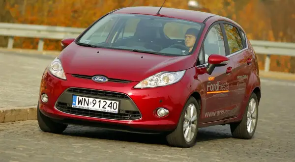 Czerwony Ford Fiesta 2010 z kierowcą za kierownicą, jadący po drodze.