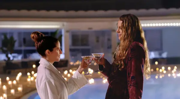 Zwei Frauen stoßen mit Martinis an, umgeben von Kerzenlicht und einem Pool. Nur noch ein kleiner gefallen, bevor der Abend perfekt ist.