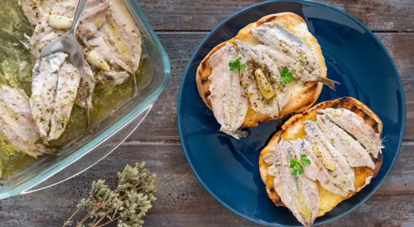 Alici al forno, profumate all'aglio e erbe, servite su crostini tostati e in una pirofila di vetro.