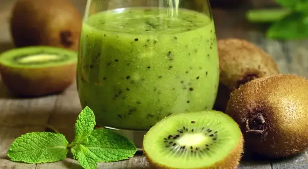 Szklanka zielonego smoothie z kiwi, pokrojone owoce kiwi i listki mięty na drewnianym tle.
