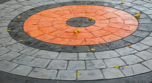 Kreisrunder Weg aus grau und orange gestrichenen Pflastersteinen, mit einigen Herbstblättern darauf.