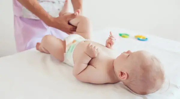 Dziecko leży na białym podłożu, a dorosły delikatnie masuje jego nóżkę. To ważny moment, wspierający ostatni skok rozwojowy malucha.