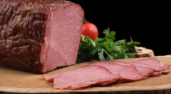 Pyszna, wędzona szynka po peklowaniu, pokrojona w cienkie plastry, z pomidorem i natką pietruszki.
