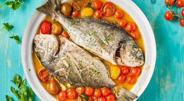 Orata all'acqua pazza, due pesci interi cotti in un brodo saporito con pomodorini e olive, guarniti con prezzemolo fresco.