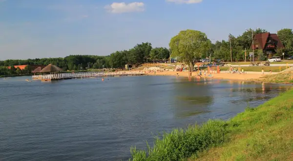 Słoneczny dzień nad jeziorem w Siemiatyczach. Plaża pełna ludzi, pomost z altaną, a w tle las i budynki. Idealne siemiatycze atrakcje.