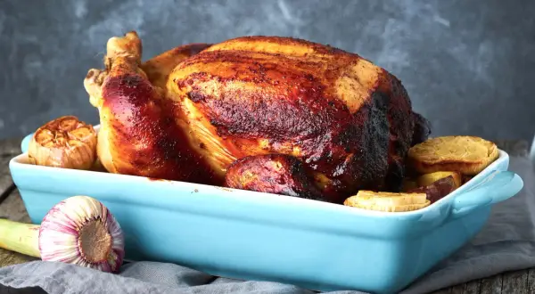Un succulento pollo al forno dorato e croccante, servito in una teglia azzurra con aglio arrosto e patate.