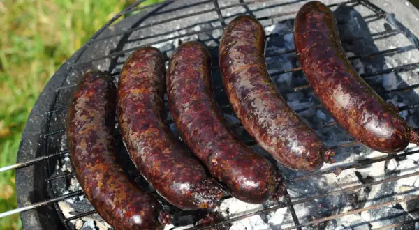 Pięć ciemnych, rumianych kiełbasek na ruszcie grilla. Widać dymek i żar, co sugeruje, jak powstaje kaszanka.