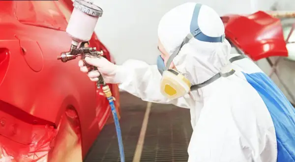 Persona in tuta protettiva e maschera effettua verniciare auto fai da te con pistola a spruzzo su un'auto rossa.