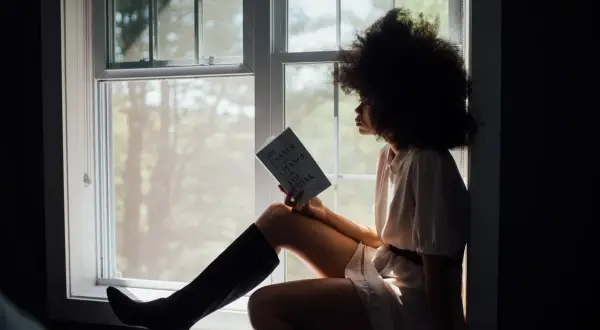 Vrouw met afro, zittend bij het raam met een boek, diep nadenkend over haar relatie.