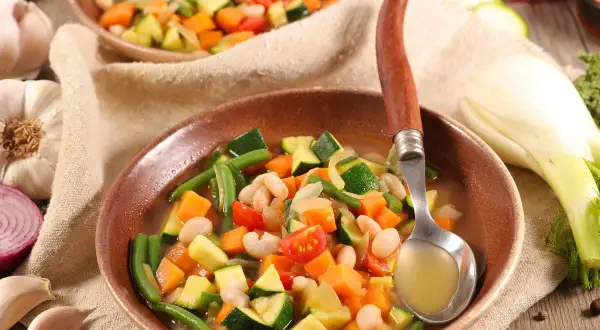 Un plato humeante de menestra de verduras, con judías verdes, zanahoria, calabacín y alubias blancas, servido con una cuchara.