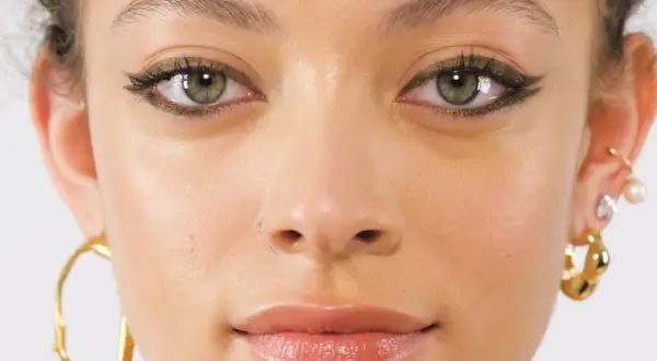 Primo piano di una ragazza con un trucco occhi audace e gioielli dorati. I suoi occhi verdi marroni sono evidenziati da un eyeliner scuro e scintillante.