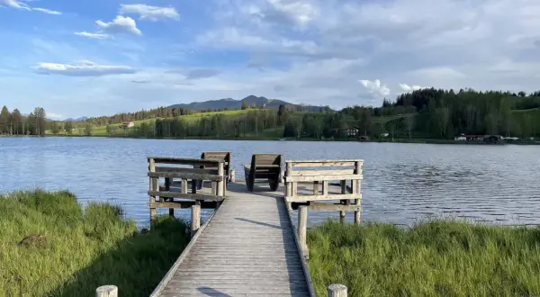 Holzsteg mit Sitzgelegenheiten am bayerischen See, umgeben von grünen Wiesen und bewaldeten Hügeln unter blauem Himmel.