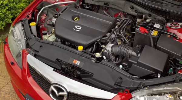 Silnik Mazdy 6 GG, czarny plastikowy kolektor dolotowy z logo Mazda, akumulator i elementy układu chłodzenia.