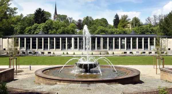 Ein Brunnen sprudelt vor einem modernen Gebäude mit Säulen und Glasfronten. Im Hintergrund ein Kirchturm und Bäume. Ein idyllischer tschechischer Kurort.