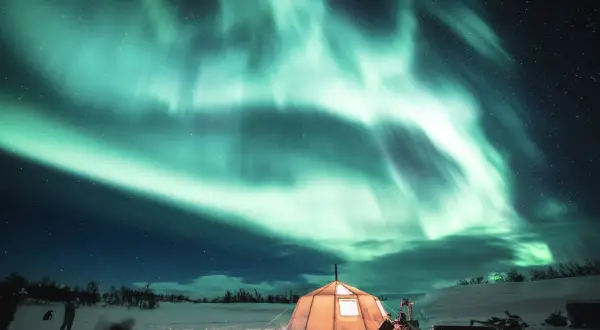 Ein beleuchtetes Zelt unter dem Nordlicht in verschneiter Landschaft.
