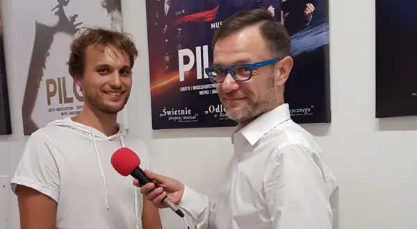 Mężczyzna w białej koszuli z niebieskimi okularami trzyma mikrofon, udziela wywiadu młodemu, uśmiechniętemu czarneckiemu aktorowi na tle plakatów teatralnych.