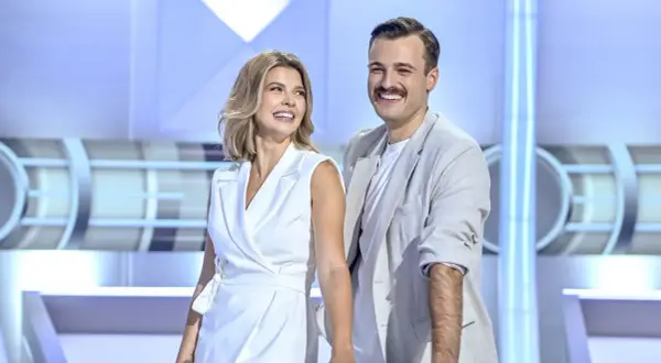Uśmiechnięci prowadzący koło fortuny, kobieta w białej sukience i mężczyzna w jasnej marynarce, stoją przy stole w studiu.