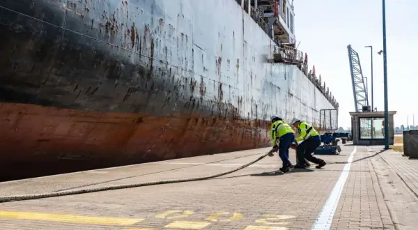Zwei Hafenarbeiter ziehen ein dickes Seil am Antwerpen Port. Ein riesiges Schiff liegt am Kai.