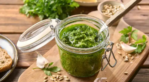 Słoik domowego pesto z pietruszki, obok czosnek, nasiona słonecznika i kromka chleba.